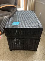 Rectangular black wicker storage trunk shown closed from an angled side view on carpeted floor next to a chair.