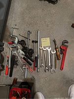 Photo showing assortment of metal hand tools and wrenches arranged on a concrete floor, including adjustable wrenches, open-end wrenches, pipe wrench, and clamping tools.