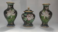 Front view of two cloisonne vases and one matching lidded pot showing floral and lotus designs on black backgrounds with gold rims and accents.
