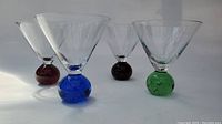 Four clear martini glasses each with a different multi-coloured spherical stem in red, blue, amber, and green, standing upright showing detail of glass design.