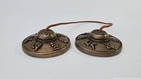 Side view of the pair of copper Tingsha cymbals connected by a leather strap, showing raised center knob and embossed characters.