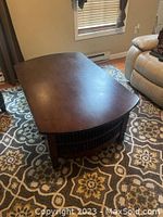 Full view of the dark brown coffee table on a patterned rug between a beige recliner and a window with blinds.
