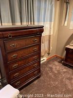 Photo of a tall, dark wood dresser with five drawers and decorative metal pulls, positioned near a window with curtains.