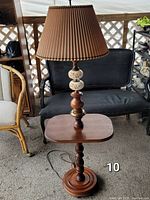 Mid century walnut floor lamp with built-in rectangular table and pleated lampshade shown in outdoor setting with surrounding furniture.