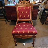 Front view of the Victorian Eastlake walnut side chair showing carved crest rail, patterned red and gold upholstery, turned front legs with casters.