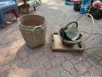 Four woven items displayed outside on paving stones: two large woven baskets with handles, one smaller woven basket with a liner and dark rim, and a rectangular woven tray with handles.