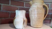 Two ceramic pitchers side by side on a flat surface against a brick wall background. Smaller white and brown pitcher on left, larger beige speckled pitcher on right.