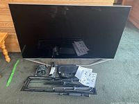 Samsung flat-screen TV, with visible reflective screen and silver-tone base stand, shown standing on carpet next to a wooden side table