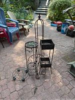 Four black metal plant stands of varying height and style with decorative scrolls and leaf accents shown together outside.