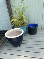 View of three pots: turquoise glazed ceramic pot with a fern plant, navy blue ceramic pot with white rim empty, and black plastic pot with blue interior empty on a wooden deck.