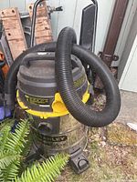 Stanley Fat Max Wet/Dry Vacuum with attached flexible black hose, showing the overall vacuum unit sitting outdoors on grass near plants, with focus on top and side view.