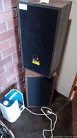 Two EPI Model 100V speakers stacked corner near a brick wall next to a white plastic humidifier with power cord.