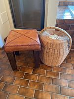 Overall view of the wood stool with leather top next to the large wicker basket with lid