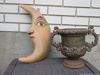 Vintage crescent moon face ornament and small cast iron urn shown side by side, illustrating their size and condition.