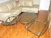 Two brass and glass tables: an oval coffee table and a rectangular side table next to a beige sectional sofa on wood floor.