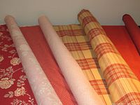 Six rolls of fabric shown together stacked horizontally, variety of red, gold, peach colors and patterns including floral, tartan, textured squares, striped velvet, and sheer fabric.