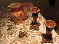 Photo showing two white resin candle holders, two metal candle holders with square bases, and large ceramic urn with floral design on a crocheted tablecloth.