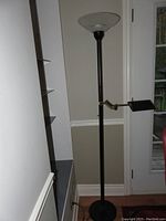 Full view of the black metal floor lamp with adjustable reading lamp arm, positioned by a white wall and a glass door.