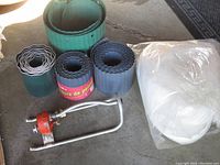 Photo of various rolls of lawn edging in black and green, a rolled white row cover fabric sealed in plastic, and a red and silver lawn sprinkler with a metal frame on concrete floor.