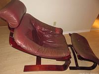 Side angled view of burgundy leather chair showing worn and torn seat and discolored leather, wooden frame base and ottoman with similar wear.