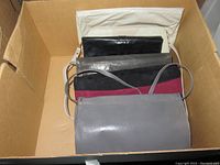 All six clutch bags arranged in a cardboard box, showing various colors and styles including gray, black, burgundy, and dark gray bags.