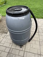 Full view of grey plastic rain barrel with black lid on paved surface and attached black overflow hose.