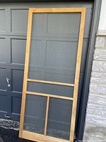 Front view of the wooden screen door resting against a garage door and wall, showing its full height, width, and mesh panel construction.