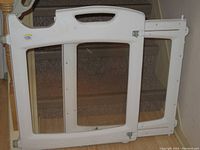 Photo of one white plastic The First Years baby gate with handle, expandable side, and transparent panel, shown standing next to carpeted stairs