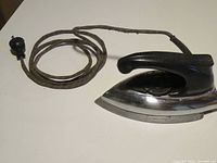 Full view of vintage electric iron with black handle, polished metal soleplate and attached cloth-covered power cord with fraying.