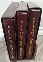 Spine view of three burgundy vinyl binders with “First Day Cover” text in gold foil