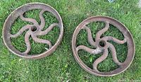 Photo of two antique cast iron wheels with curved spokes lying on grass, showing overall shape, rust, and size.