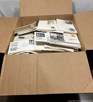 Open cardboard box filled with a large assortment of First Day Covers and Postal Covers with various stamps and cancel marks.