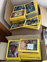 Two cardboard boxes filled with stacks of National Geographic magazines showing the yellow bordered covers with various images, including animals, the Statue of Liberty, and people.