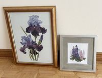 Two framed needlepoint artworks on floor side by side, one larger with purple irises in a gold frame, one smaller with lupin flowers in a silver frame.
