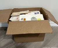 Wide angle view of large cardboard box filled with assorted first day covers and postal covers showing variety of stamps, some with colorful designs