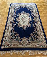 Full view of the vintage hand hooked, hand carved area rug laid on parquet floor showing royal blue background and floral border design with fringe.