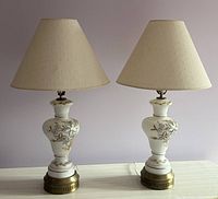 Pair of vintage table lamps with frosted white glass, brass bases, and fabric lamp shades standing side by side on a white surface against a pale purple background.