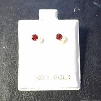 Front view of the pair of white gold stud earrings with red garnet stones on a white holder marked 10K T. GOLD