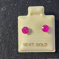 Front view of the pair of round ruby gemstone stud earrings set in 10K white gold four-prong settings on a white card marked 10 KT. GOLD