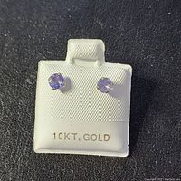 Front and angled view of two round-cut tanzanite stud earrings set in 10K yellow gold on a white textured holder labeled '10KT. GOLD'.