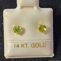 Close-up front view of two heart-shaped peridot gemstones set in 14K yellow gold stud earrings on a 14K gold marked display card.