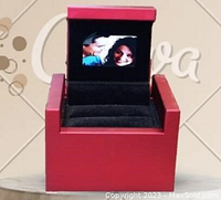 Red rectangular jewelry box with built-in screen showing video of a couple, lid open, black velvet interior.