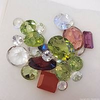 Distinct loose faceted gemstones in multiple colors including green, purple, blue, red, orange, and clear displayed on a white background.