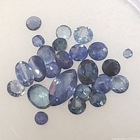 Close-up photo showing multiple faceted blue sapphire gemstones in various shapes and shades, positioned on white foam inside a container.