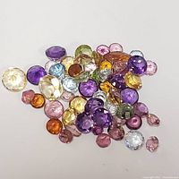 Photo showing a loose assortment of mixed colored faceted gemstones on a white surface, including purple, yellow, orange, green, and pink stones.