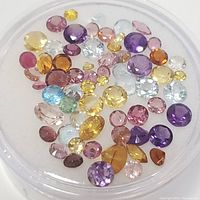Photo showing assortment of loose faceted multicolor gemstones in a small clear round container. Various colors visible include yellow, purple, pink, blue, green, red and clear. Mostly round cuts with some marquise shaped stones.