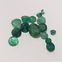 Photo of fourteen loose genuine emerald gemstones showing different sizes and cuts on a white background.