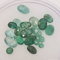 Top view of 21 oval-shaped emerald gemstones in a clear container showing various shades of green and visible inclusions.
