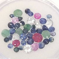 Top-down image of assortment of polished, faceted loose gemstones including green emeralds, red rubies, blue sapphires, pink stones, and clear stones in various sizes placed on a white background.