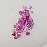 Photo shows an assortment of small round and faceted pink sapphire gemstones scattered on a light surface, displaying their pink hues and clarity.